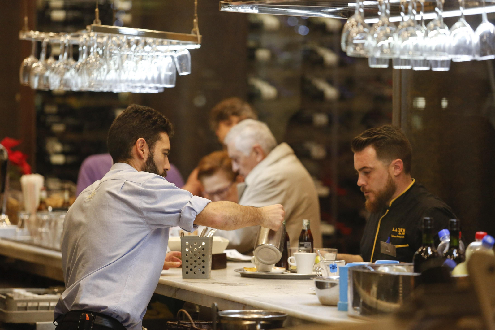Un trabajador desarrolla sus labores en la barra de un bar en el que hay varios clientes.